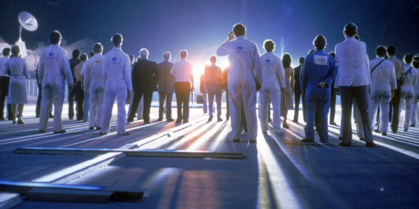 Silhouetted crowd of actors in jumpsuits looking toward a bright light source on a landing pad at night; iconic scene from Close Encounters of the Third Kind.