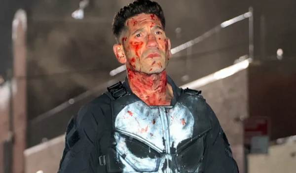 Jon Bernthal as Frank Castle in Daredevil: Born Again, spattered in blood and wearing a tactical vest with the iconic white Punisher skull spray-painted on the chest. He is standing in a dark, gritty indoor setting.