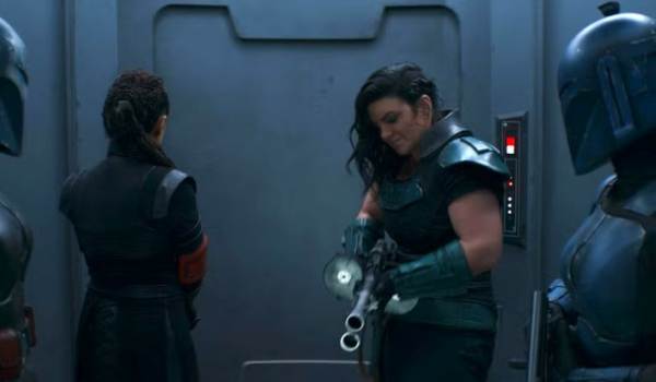 A scene from The Mandalorian featuring Cara Dune (Gina Carano) checking a large heavy blaster while standing between two Mandalorians in blue armor and Fennec Shand. They are positioned in a metallic, dimly lit hallway.