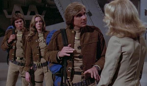 A still from the original Battlestar Galactica series showing Captain Apollo (Richard Hatch), Lieutenant Starbuck (Dirk Benedict), and Cassiopeia (Maren Jensen) in their tan Colonial uniforms, talking to a blonde woman.