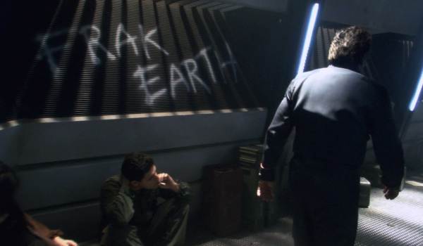 : A gritty scene from the Battlestar Galactica reimagined series showing the words "FRAK EARTH" spray-painted on a wall inside the ship, with a crew member sitting nearby.