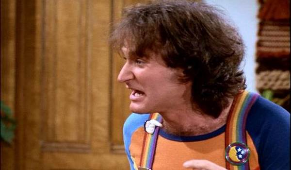 Robin Williams as Mork in Mork & Mindy, wearing his signature rainbow suspenders and a colorful t-shirt while speaking with an animated expression.