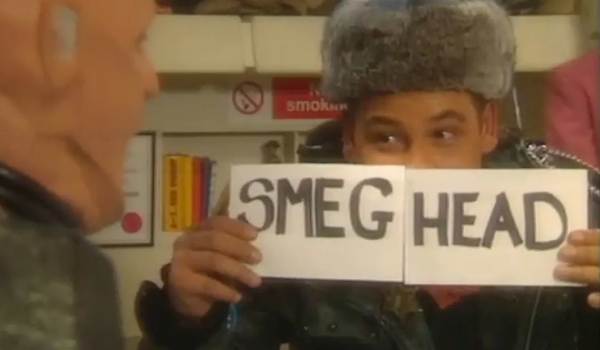 A scene from the sci-fi comedy Red Dwarf showing Dave Lister (Craig Charles) wearing a fur hat and holding up signs that read "SMEG HEAD" while looking at the mechanoid Kryten.