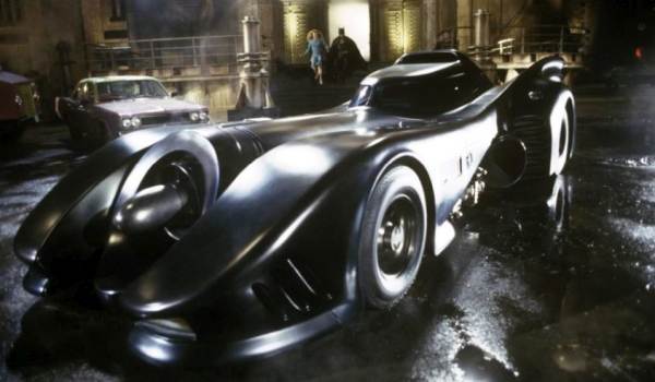 Frontal three-quarter view of the 1989 Batmobile parked outside the Flugelheim Museum with Batman and Vicki Vale in the background.