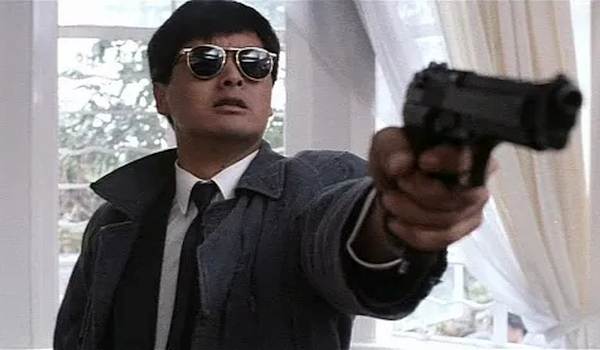Chow Yun-fat as Mark Gor in A Better Tomorrow (1986), wearing sunglasses and a trench coat while pointing a handgun during an action sequence.