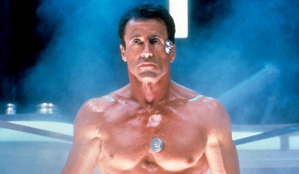 Sylvester Stallone as John Spartan in Demolition Man, shown shirtless with futuristic sensors attached to his chest and face against a glowing blue background.