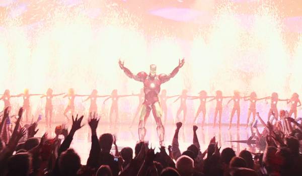 Tony Stark (Robert Downey Jr.) stands with arms outstretched as pyrotechnics erupt behind him during the Stark Expo in Iron Man 2. The Ironettes dance in a line behind him while a cheering crowd fills the foreground.