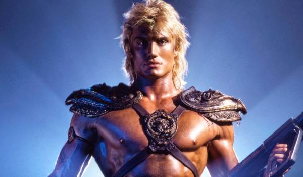 Dolph Lundgren as He-Man in the 1987 film Masters of the Universe, wearing leather harness armor and shoulder pauldrons while holding a weapon against a bright blue background.