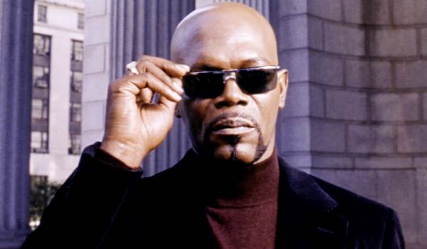Samuel L. Jackson as John Shaft in Shaft (2000), wearing a brown turtleneck and black coat while adjusting his sunglasses in front of a stone building.