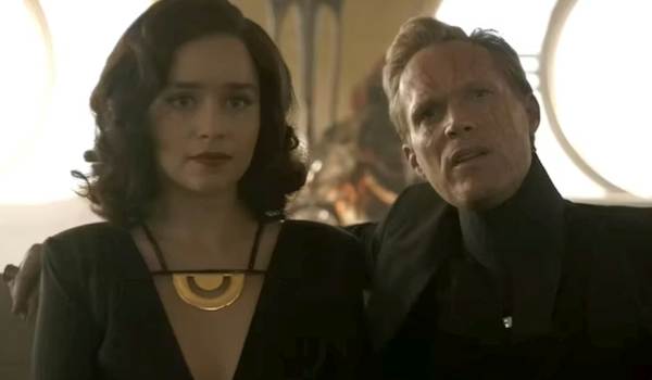 Qi'ra and Dryden Vos sitting together aboard a luxury yacht in a scene from Solo: A Star Wars Story. Qi'ra wears a black gown with a prominent gold necklace, while Dryden Vos, showing visible facial scars, looks on with a stern expression.