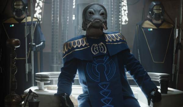 An image from The Book of Boba Fett ("Chapter 7: In the Name of Honor") showing the Pyke Boss leaning over a white table. He is a grey-skinned humanoid with large dark eyes, wearing ornate blue and gold robes. Two armored sentinels stand behind him in the background.