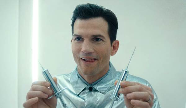 Ashton Kutcher as Byron Forst in the FX series The Beauty, wearing a metallic silver jacket and holding two large syringes in a sterile white lab, featuring a mischievous and intense expression.