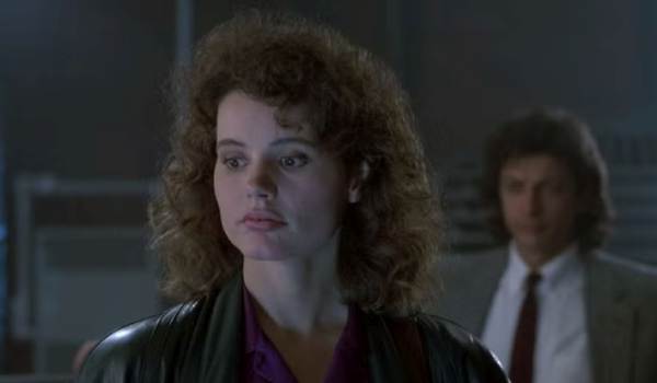 Geena Davis as Veronica Quaife and a blurred Jeff Goldblum as Seth Brundle in the 1986 film The Fly, featuring Davis with voluminous curly hair and a leather jacket.