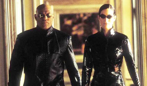 Laurence Fishburne as Morpheus in a long crocodile-skin coat and Carrie-Anne Moss as Trinity in a shiny PVC bodysuit walking side-by-side through a hallway in The Matrix.