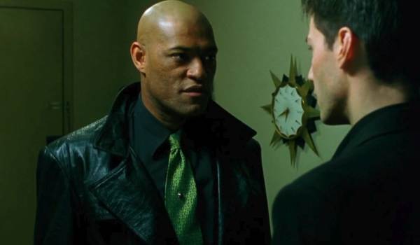 Laurence Fishburne as Morpheus wearing a black leather coat and green tie, speaking with Neo (Keanu Reeves) in a room with a starburst clock in The Matrix (1999).