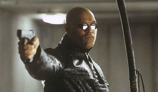 Laurence Fishburne as Morpheus in The Matrix Reloaded (2003), wearing a textured black leather coat and sunglasses while wielding a katana and pointing a handgun.