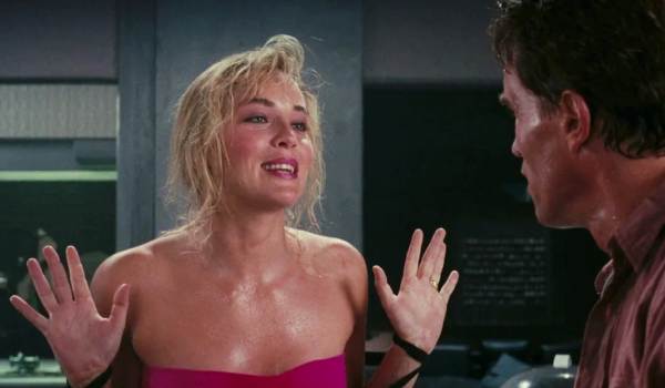 Lori Quaid (Sharon Stone) holds her hands up with a deceptive smile while facing Douglas Quaid (Arnold Schwarzenegger) in a scene from the 1990 film Total Recall. Lori wears a pink athletic top and appears disheveled after a fight, set against the futuristic interior of their apartment.