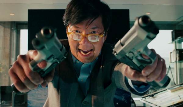 Ken Jeong as Jerry Wang in Transformers: Dark of the Moon, wearing yellow-rimmed glasses and a grey suit while holding two silver handguns toward the camera with an intense, shouting expression.