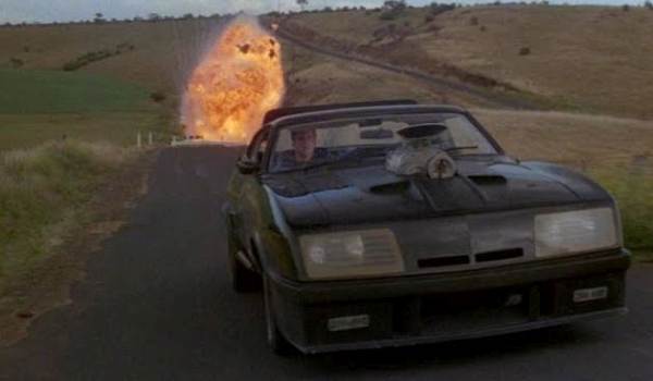 Frontal view of the V8 Interceptor driving away from a large explosion on a desert road in The Road Warrior.