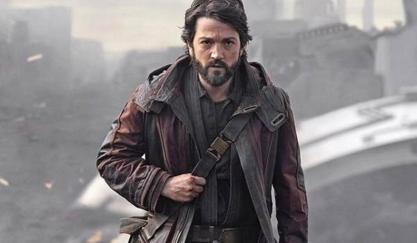 Cassian Andor (Diego Luna) looking intense and determined while wearing a heavy brown coat and a messenger bag, standing amidst the industrial ruins of Ferrix in the 2022 series Andor.