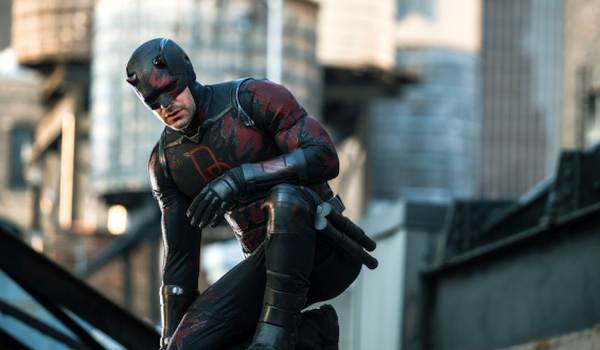 Charlie Cox as Daredevil kneels on a rooftop in a black and red tactical suit, looking downward with a focused expression.