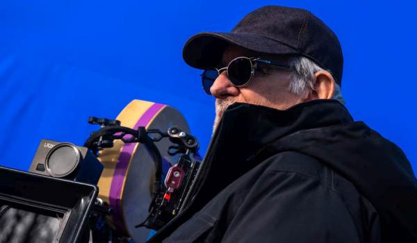 Close-up of Steven Spielberg wearing a cap and sunglasses while filming a scene for Disclosure Day in front of a large production blue screen.