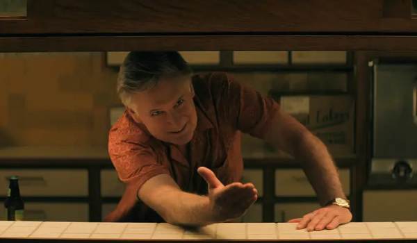 Actor Bill Pullman as Jack leans forward through a kitchen pass-through counter, gesturing with an open hand in a scene from Netflix's The Boroughs.
