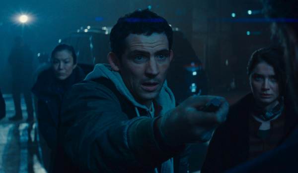 A close-up from Disclosure Day shows a wide-eyed Dr. Daniel Kellner (Josh O'Connor) in a hoodie and dark jacket, holding out a small object in his right hand. He is flanked by Hettienne Park as Serena and Eve Hewson as Jane Blankenship in a dimly lit, blue-toned outdoor setting at night.