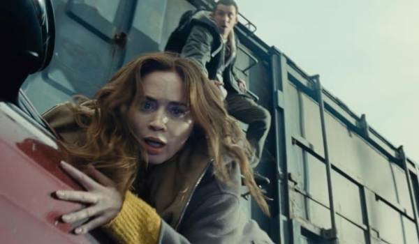 An intense, low-angle shot shows a terrified Margaret Fairchild (Emily Blunt) clinging to the side of a red vehicle, while Dr. Daniel Kellner (Josh O'Connor) looks on anxiously from a ladder on a moving cargo train in the background of the sci-fi film Disclosure Day.