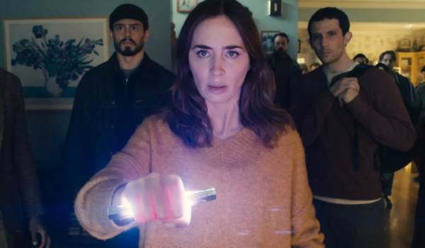 A medium close-up shows a wide-eyed Margaret Fairchild (Emily Blunt) in a tan sweater holding a glowing cylinder toward the camera, flanked by Santiago (Tommy Martinez) and Dr. Daniel Kellner (Josh O'Connor) in a dimly lit room.