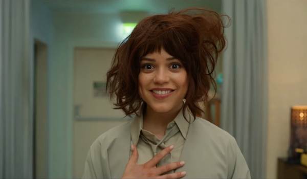 Jenna Ortega as Klara in the movie Klara and the Sun, smiling broadly with one hand on her chest while wearing a messy brown wig and a light-colored collared uniform.