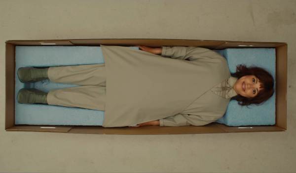 An overhead, bird's-eye view of Jenna Ortega as Klara in the movie Klara and the Sun, lying perfectly still and smiling inside a long cardboard shipping box lined with blue foam inserts.