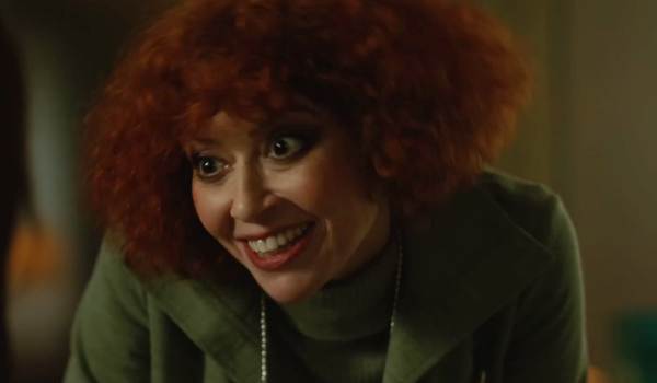 Natasha Lyonne in the movie Klara and the Sun, smiling with a wide, intense expression while wearing a green coat and a turtleneck