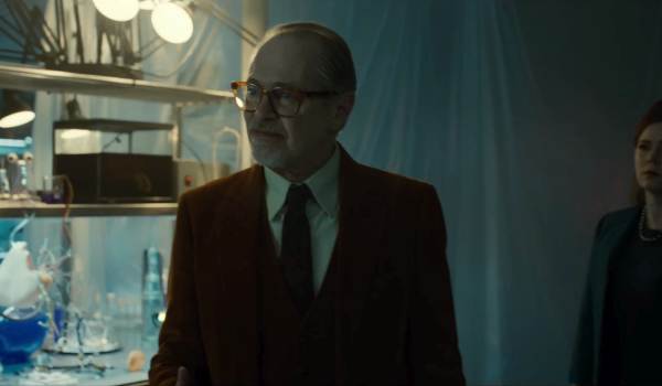 Steve Buscemi in the movie Klara and the Sun, wearing a brown suit and glasses while standing in a dimly lit, laboratory-like setting
