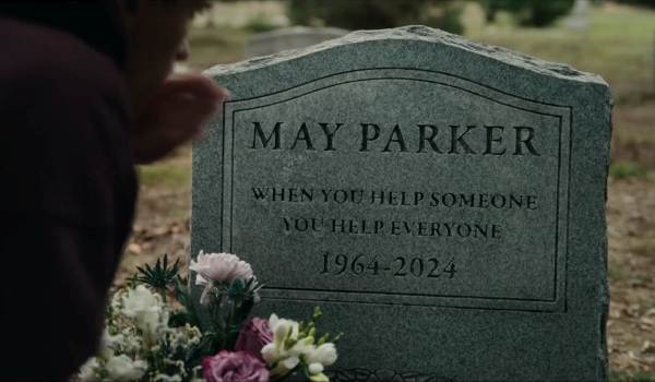 A close-up of May Parker's tombstone reading "When you help someone you help everyone 1964-2024," with a bouquet of flowers in the foreground and a person partially visible on the left, from a scene in Spider-Man: Brand New Day.