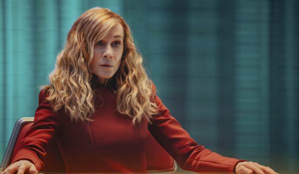 Medium shot of Holly Hunter as Captain Nahla Ake in the series Star Trek: Starfleet Academy, seated at a desk and wearing a formal red Starfleet uniform while looking intently off-camera.