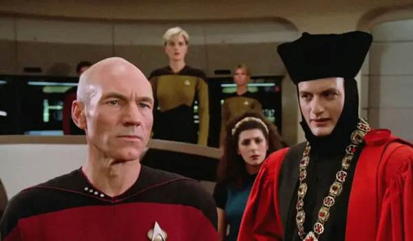 Patrick Stewart as Captain Jean-Luc Picard standing sternly alongside John de Lancie as Q, who is dressed in a red and black judge's robe, with Marina Sirtis as Counselor Deanna Troi and Denise Crosby as Lieutenant Natasha 'Tasha' Yar on the bridge of the USS Enterprise in the sci-fi show Star Trek: The Next Generation.