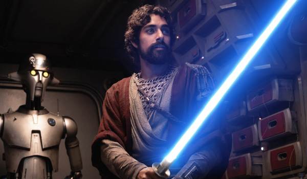 A live-action shot from the Ahsoka series showing a bearded Ezra Bridger, played by Eman Esfandi, in textured maroon robes holding an ignited blue lightsaber, with the droid Huyang, voiced by David Tennant, standing behind him