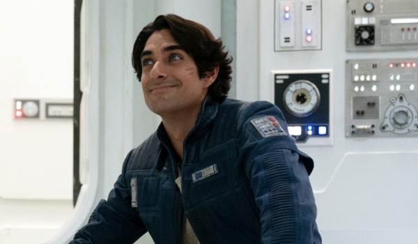 A live-action shot from the Ahsoka series featuring a clean-shaven Ezra Bridger, played by Eman Esfandi. He is wearing a dark blue New Republic pilot-style jacket and looking upward with a warm, nostalgic smile inside a bright white starship corridor.