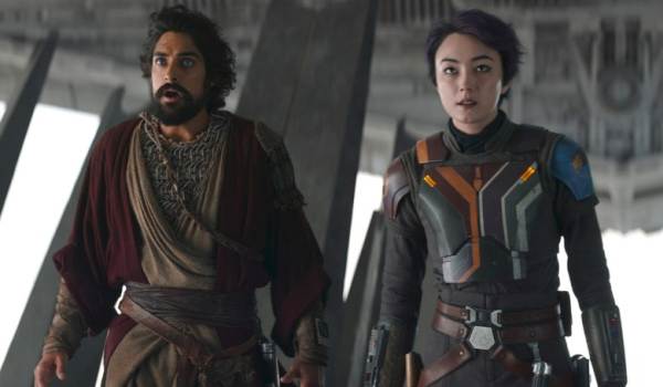 A live-action shot from the Ahsoka series showing a wide-eyed Ezra Bridger, played by Eman Esfandi, and a focused Sabine Wren, played by Natasha Liu Bordizzo, standing side by side in full combat gear inside an industrial hanger bay.