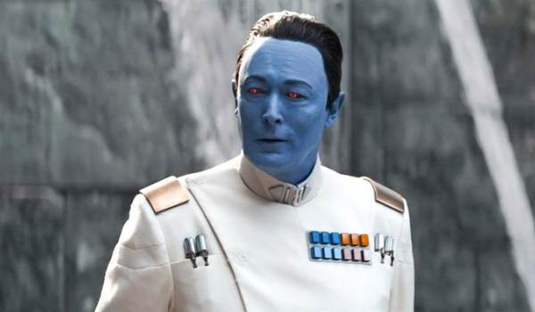 A close-up live-action shot from the Ahsoka series featuring Grand Admiral Thrawn, played by Lars Mikkelsen. He has blue skin, striking red eyes, slicked-back dark hair, and wears a white Imperial Grand Admiral uniform with a multi-colored rank insignia plaque