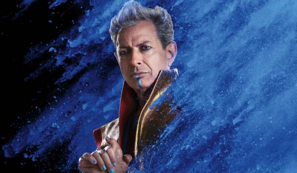 Promotional character poster of Jeff Goldblum as the Grandmaster from the 2017 movie Thor: Ragnarok, featuring blue facial makeup and blue fingernails against a disintegrating blue dust background.
